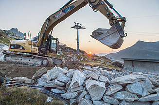 Crawler excavator operating on a construction site in alpine mountain terrain with rock material, infrastructure buildings and a cable car installation in the background.