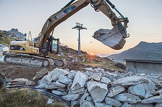 Crawler excavator operating on a construction site in alpine mountain terrain with rock material, infrastructure buildings and a cable car installation in the background.