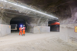 Interior view of an underground structure with multiple tunnel passages, lighting, ventilation systems and operational infrastructure. Two workers wearing protective equipment are standing near a tunnel junction.