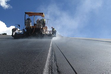  Road paving machine laying a new asphalt surface on a transport infrastructure project. The foreground shows the freshly paved asphalt layer and a longitudinal joint running across the surface.