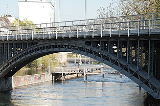 Stahlbogenbrücke mit Fahrbahn über einem Kanal in urbaner Umgebung. Sichtbar sind die genietete Stahlkonstruktion, steinerne Widerlager und das Gewässer unterhalb der Brüc
