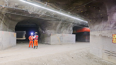 Interior view of an underground structure with multiple tunnel passages, lighting, ventilation systems and operational infrastructure. Two workers wearing protective equipment are standing near a tunnel junction.