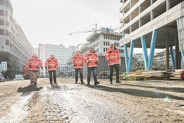 Five construction engineers can be seen walking across a building site, with new buildings in the background.