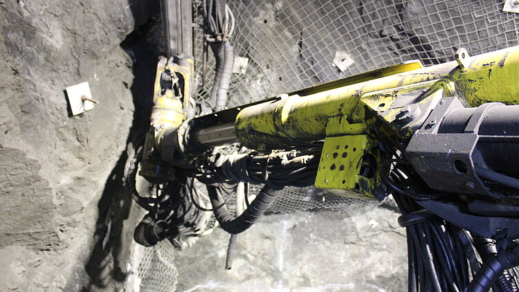 Hydraulic drilling equipment operating inside an underground mine tunnel, with rock surfaces, mining infrastructure and support systems visible along the excavation.
