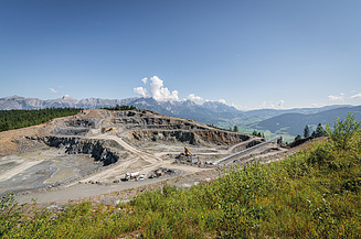 Quarry featuring terraced extraction areas, heavy machinery and conveying equipment set within an alpine landscape. Forested slopes and mountain ranges are visible in the background.