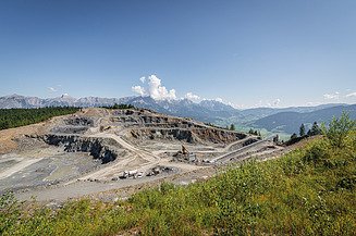 Quarry featuring terraced extraction areas, heavy machinery and conveying equipment set within an alpine landscape. Forested slopes and mountain ranges are visible in the background.