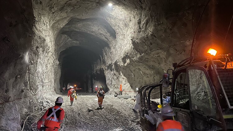 Underground tunnel at El Teniente Mine in Chile with workers in protective equipment, service vehicles, rock support systems and operational mining infrastructure visible along the excavation.