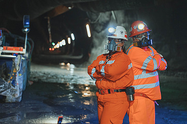 Zwei Mitarbeitende mit Schutzausrüstung in einem unterirdischen Stollen der Mine El Teniente in Chile. Im Hintergrund sind Tunnelinfrastruktur und bergbauliche Anlagen erkennbar.