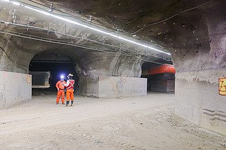 Interior view of an underground structure with multiple tunnel passages, lighting, ventilation systems and operational infrastructure. Two workers wearing protective equipment are standing near a tunnel junction.