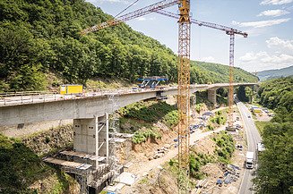 Bridge construction site in a forested valley with tower cranes, reinforced concrete bridge structures and construction areas alongside a roadway.