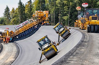 Spezialfertiger und Straßenwalzen beim Einbau und der Verdichtung einer Asphaltdeckschicht auf der geneigten Böschung einer Damm- oder Speicheranlage.