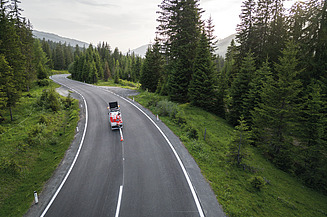 Luftbild einer asphaltierten Straße in einer bewaldeten Gebirgslandschaft mit einem Servicefahrzeug auf der Fahrbahn und umliegender alpiner Vegetation.